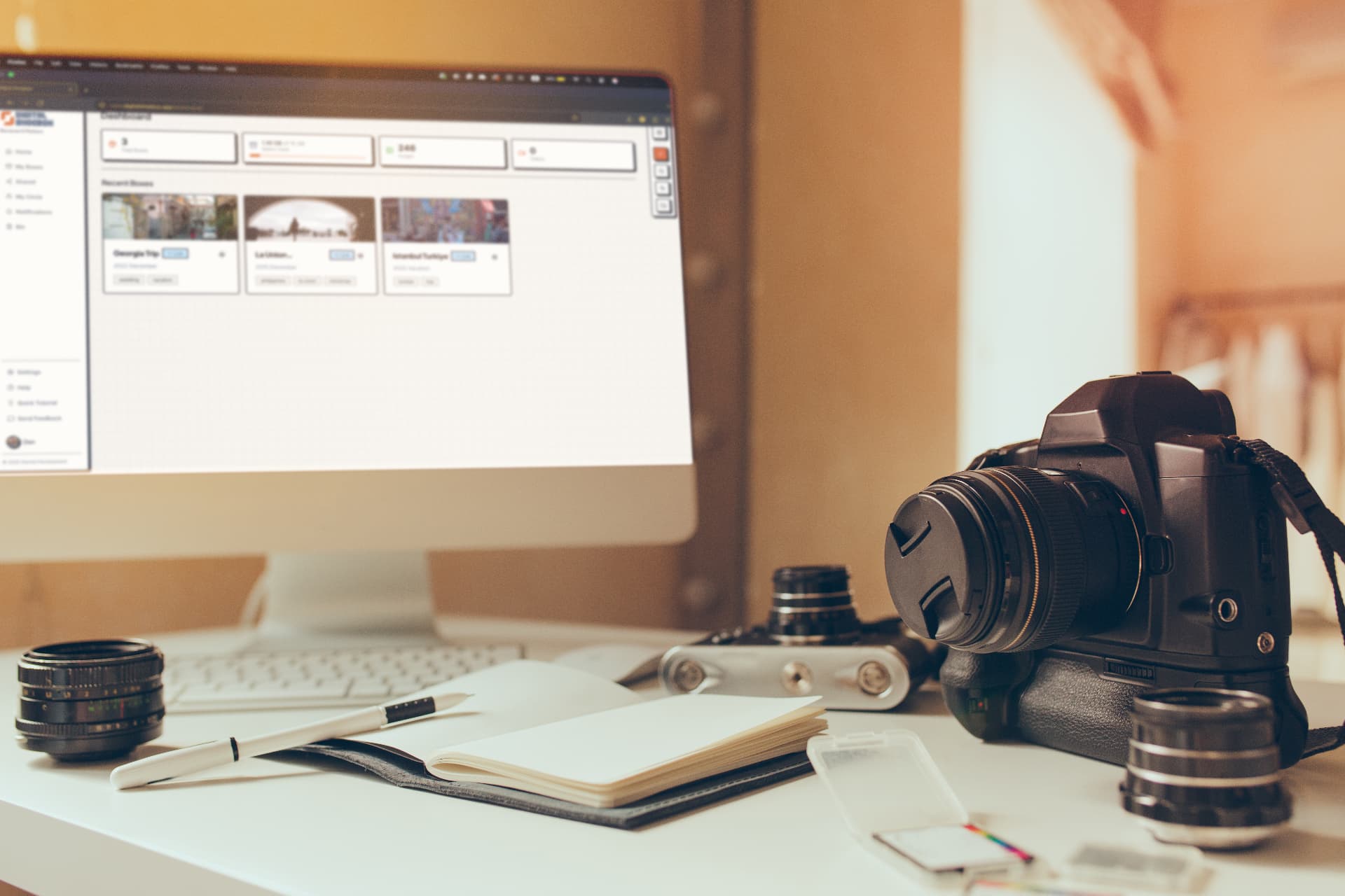 Modern iMac workstation displaying the Digital Shoebox dashboard beside a professional DSLR camera, lenses, notebook, and SD cards